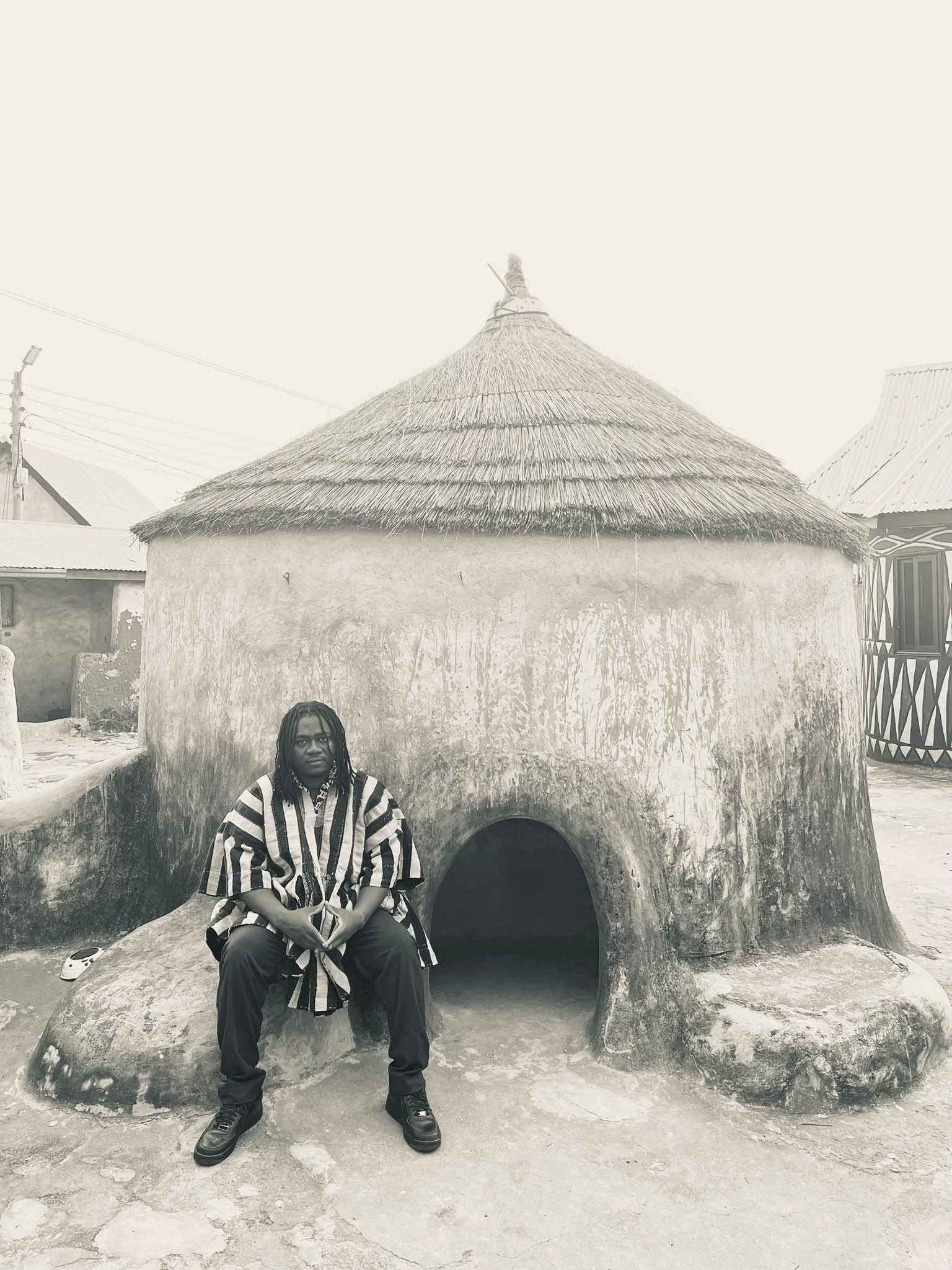 Soorebia in a striped smock seated in front of a traditional thatched building